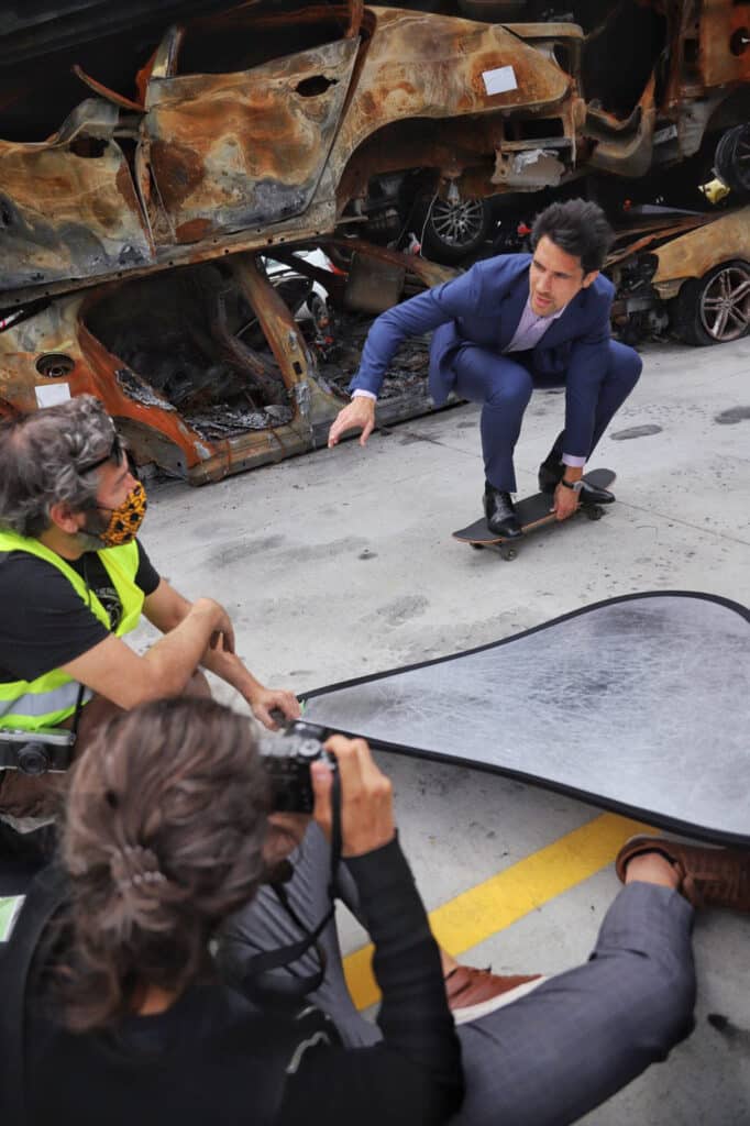 Coulisses d'un shooting photo corporate : homme en costume sur un skateboard devant des carcasses de voitures.