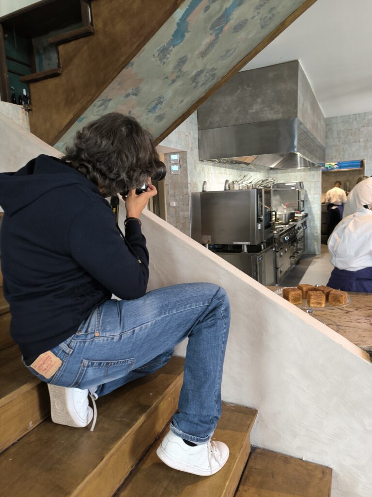 Photographe assis sur un escalier en bois, prenant une photo d’une cuisine professionnelle ouverte.