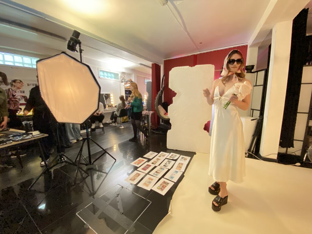Une mannequin en robe blanche et lunettes de soleil pose avec un bouquet devant un fond blanc, dans un studio animé par une équipe en coulisses.
