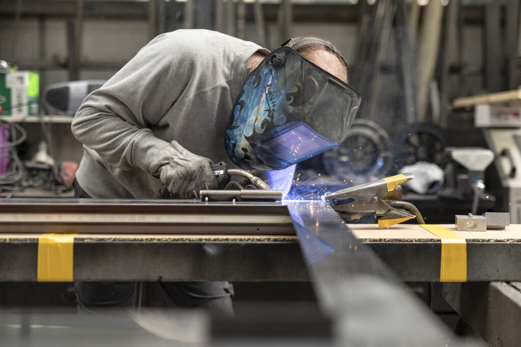 Un soudeur expert en gants et masque de protection bleu travaille sur le coin d'un profilé métallique dans un atelier industriel, avec l'arc de soudure et des étincelles visibles.