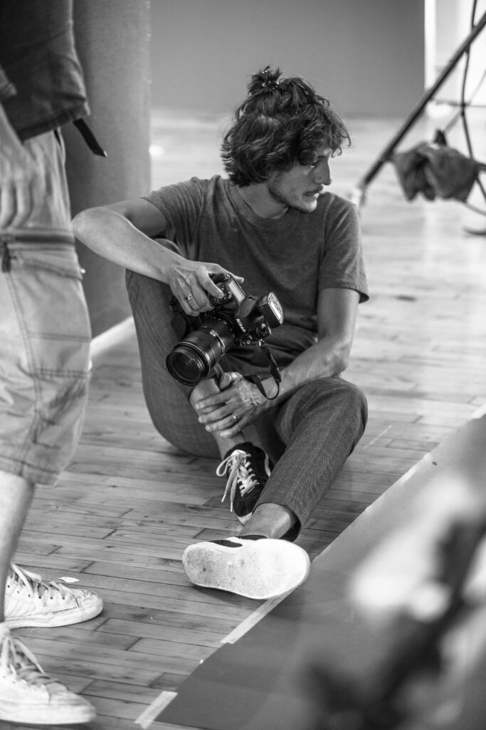 Portrait en noir et blanc d'un photographe assis au sol tenant un appareil reflex Nikon avec flash dans un studio au parquet de bois