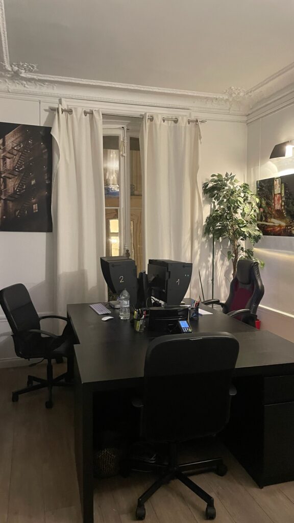 Espace de travail avec des bureaux noirs alignés, des fauteuils ergonomiques et des écrans d'ordinateur équipés de casquettes anti-reflet dans un bureau élégant avec moulures.