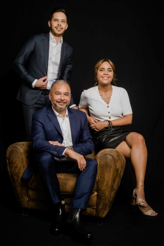photo corporate paris en studio de trois collaborateurs, un homme assis en costume bleu marine entouré de deux collègues, fond noir, Studio Alterego