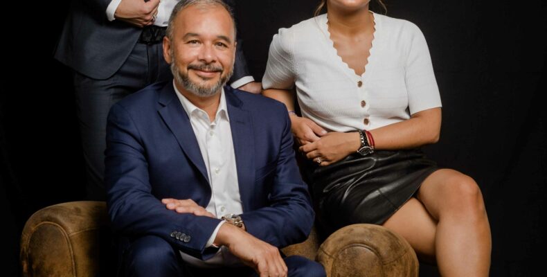 photo corporate paris en studio de trois collaborateurs, un homme assis en costume bleu marine entouré de deux collègues, fond noir, Studio Alterego